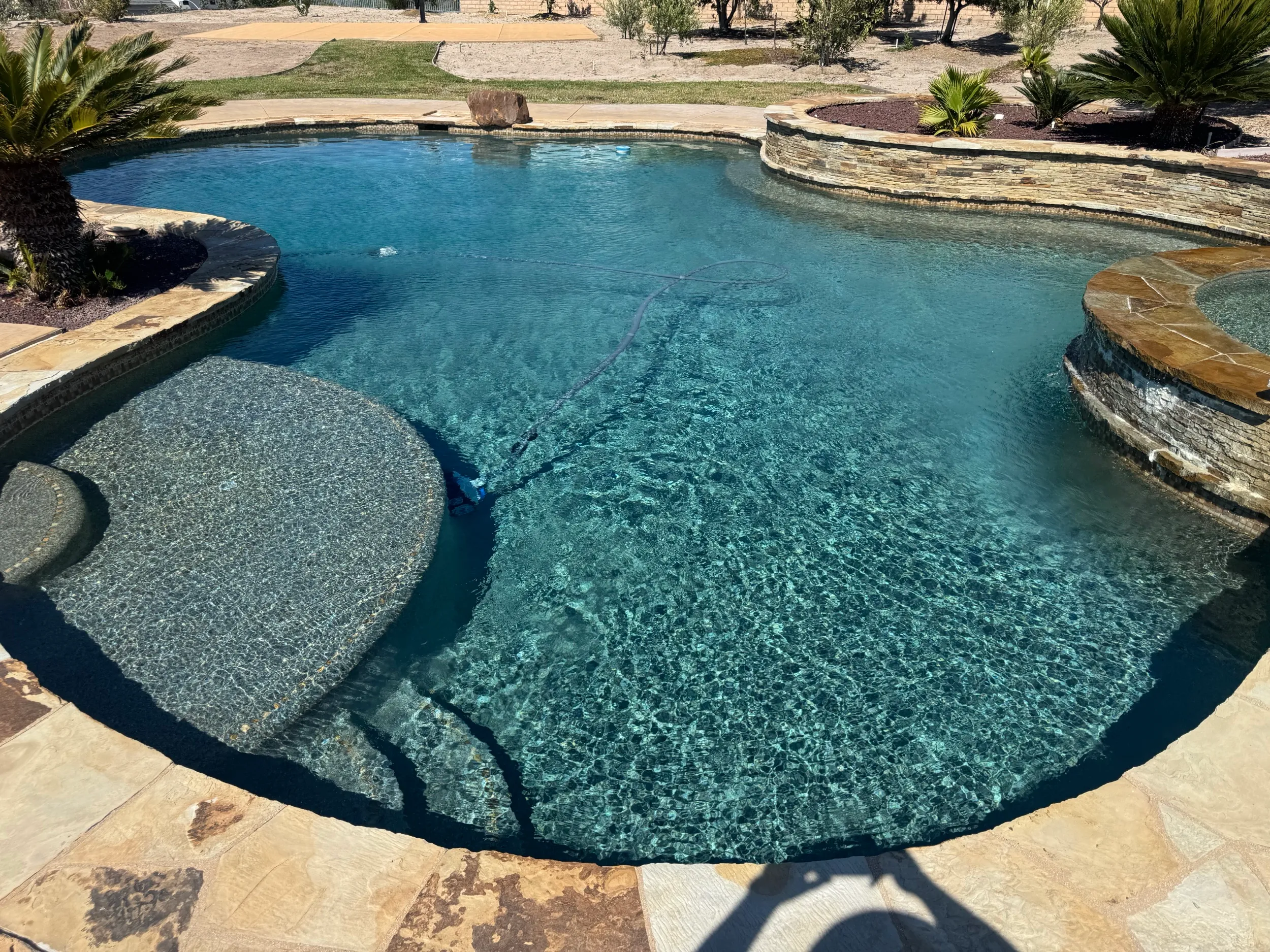 Crystal-clear backyard pool with a flagstone deck and a stone wall, with a pool cleaner at work on the bottom